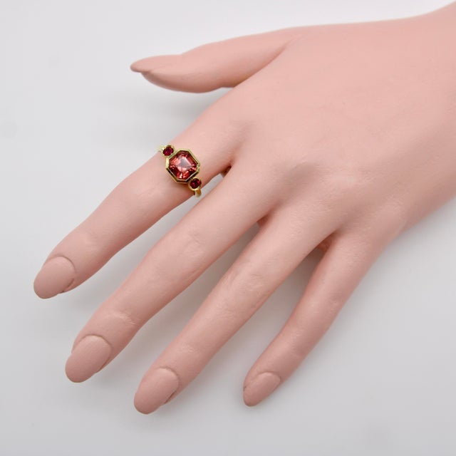 This 18k gold three stone ring features a peach tourmaline as its focal point. Sourced from Tanzania, the tourmaline is a...