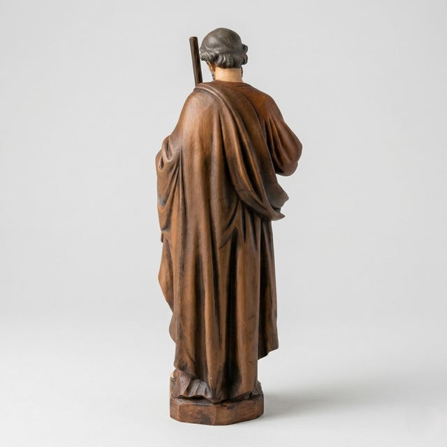 Saint Joseph is depicted standing with a staff held close to the body and one hand resting over his chest. Layered brown...