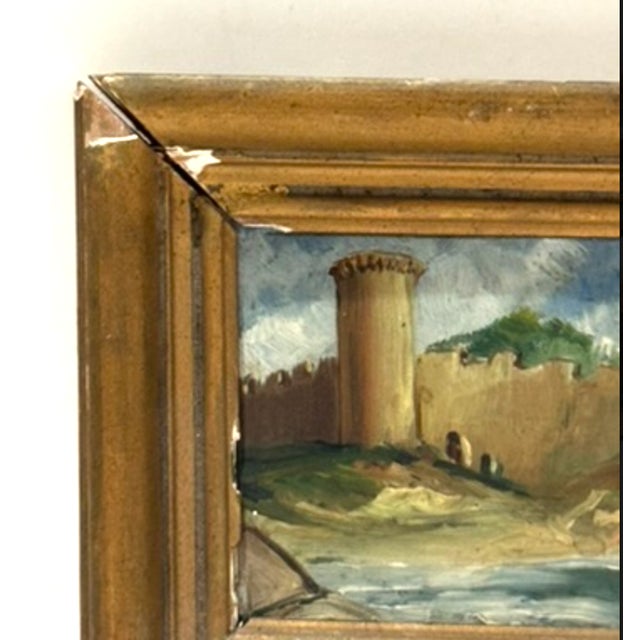 Small vintage miniature landscape painting depicting a tower and stone wall along a shoreline or riverbank under a cloudy...