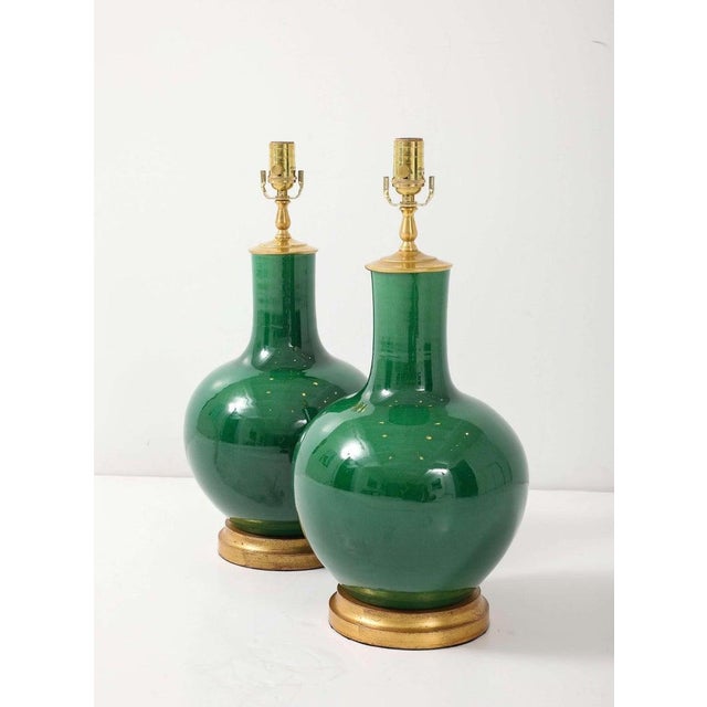 A pair of ceramic table lamps finished in a deep, high-gloss emerald glaze. Each lamp sits on a hand-gilded wood base and...