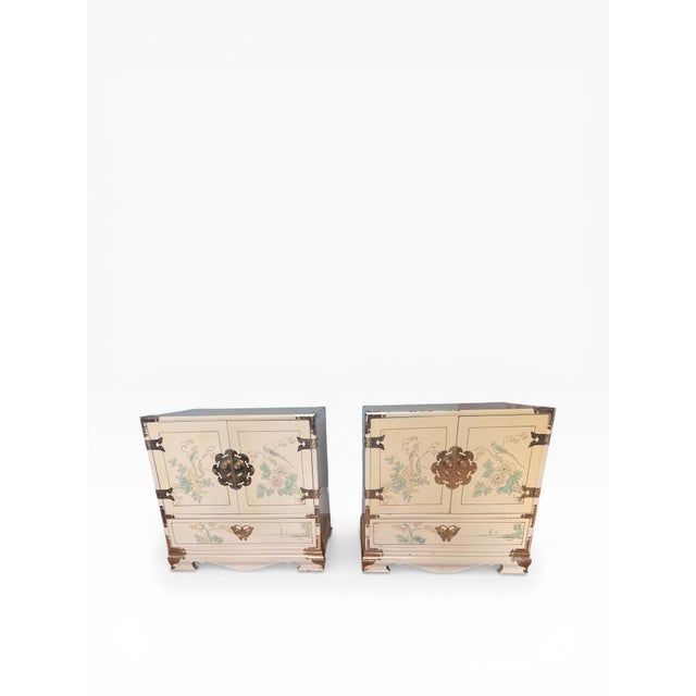 A decorative pair of vintage chinoiserie nightstands featuring a cream lacquered finish with hand-painted bird and floral...