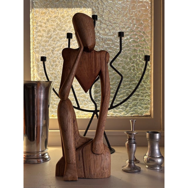 Striking hand-carved wood sculpture from Ghana depicting a stylized seated figure in a contemplative pose. The elongated...