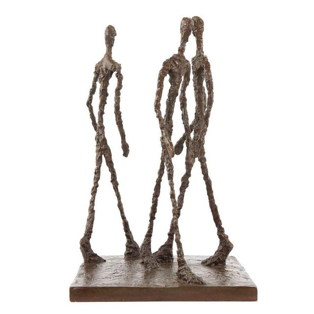 This modern dark patinated bronze sculpture is a figurative artwork depicting three elongated male figures walking...