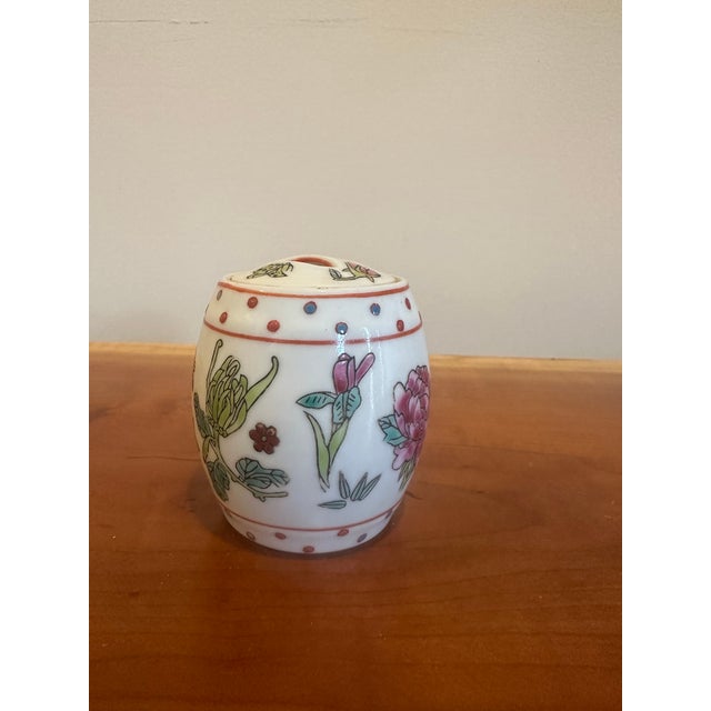 A petite porcelain jar featuring hand-painted floral motifs in soft pinks, greens, and delicate detailing. The domed lid...