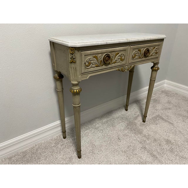 Elegant vintage Louis XVI-style console table featuring an antiqued gray-beige painted wood finish with gilt accents and...