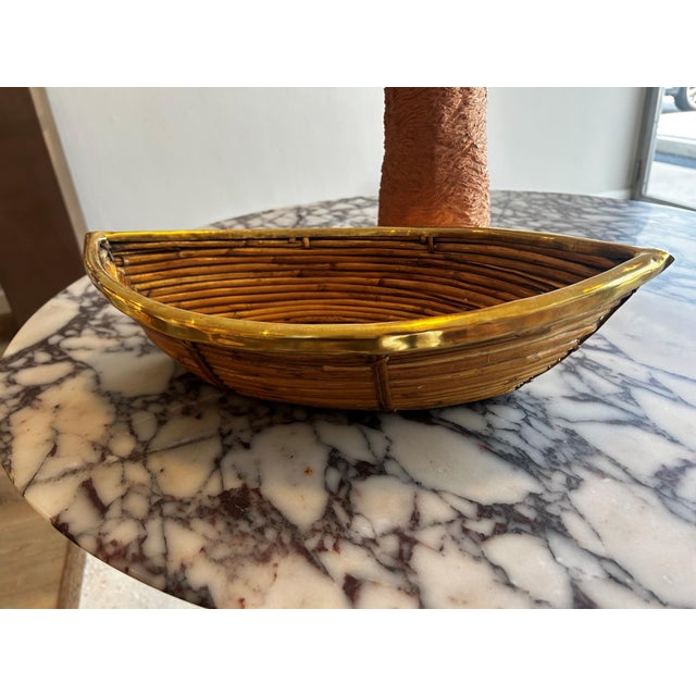 An elongated pencil reed basket with brass trim, crafted in Italy and shaped in a distinctive boat form. The woven...