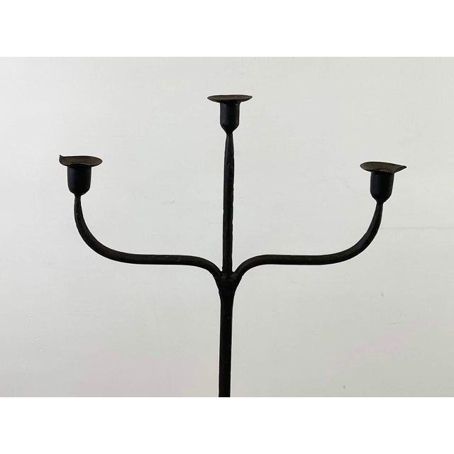 The tall, black iron candelabra features three arms supporting taper candles, leading to a twisted central shaft, raised...