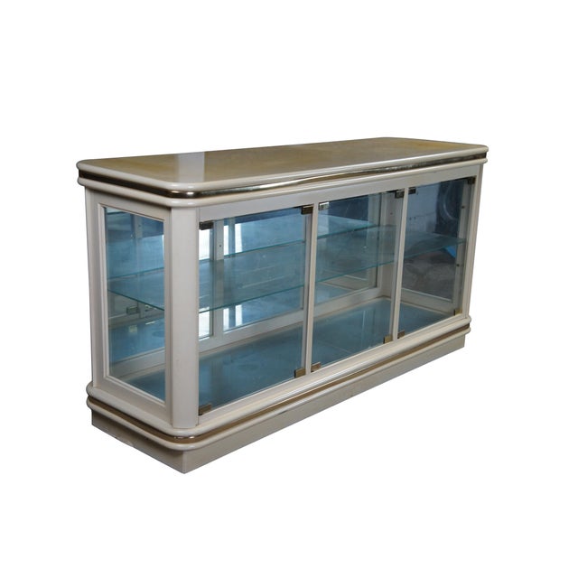 Hollywood Regency Console Cabinet or Showcase. Features and off-white finish with brass accents. Interior opens via three...