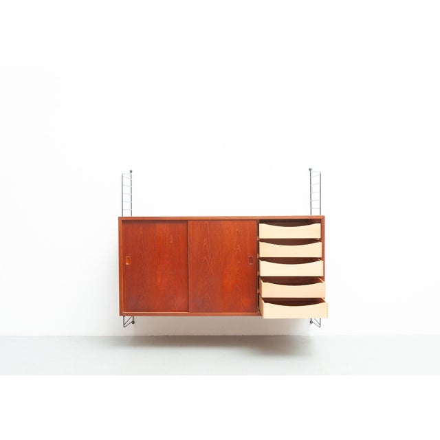 Wall-mounted teak sideboard with three sliding doors. Behind the right-hand door are five drawers. Behind the left and...