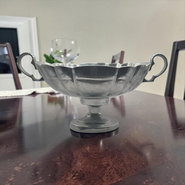 This elegant serving vessels is crafted from pewter and features a fluted body, decorative handles, and a pedestal base....
