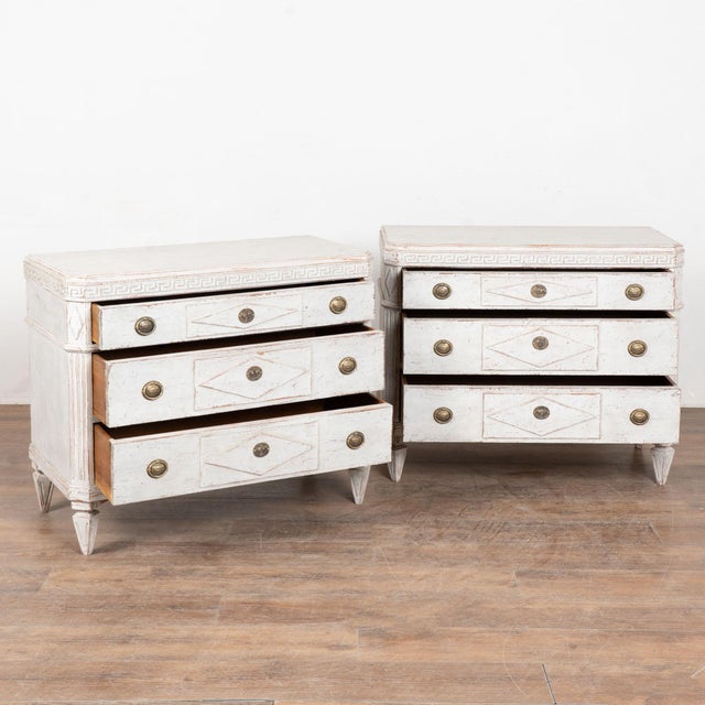 A pair of decorative Gustavian pine chest of drawers painted in shades of antique white with Greek key motif carved in...