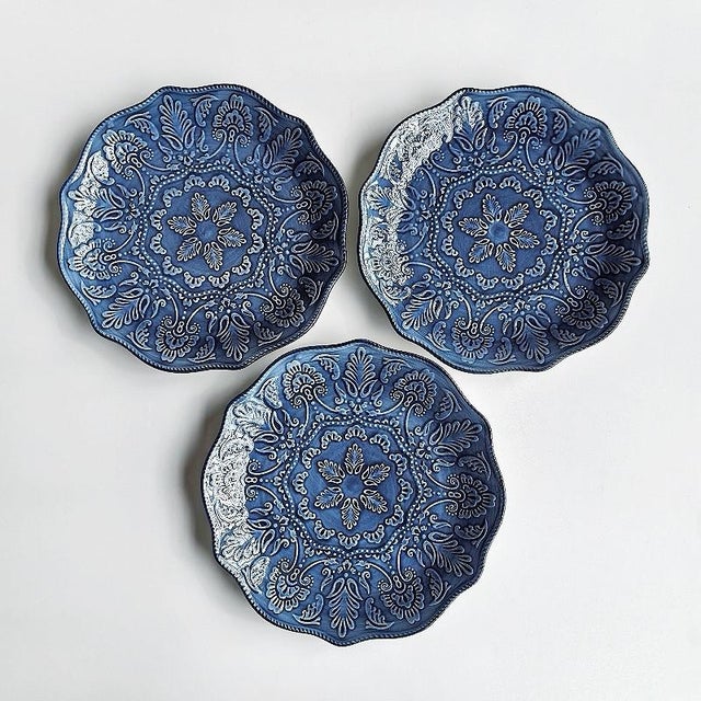 A four-piece set of blue embossed ceramic plates featuring a raised, vintage-style floral pattern. Intricate, swirling...