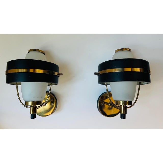 An original pair of 1950s Italian mid century sconces in golden polished brass and black enamel with white frosted glass...
