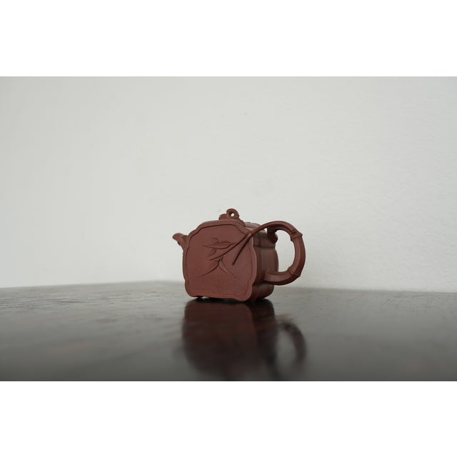 This Chinese pottery teapot features a sculptural relief design with a bamboo motif sweeping across its rounded, panel-...