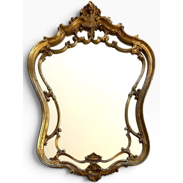 A beautifully ornate vintage wall mirror with an elegant sculptural silhouette and richly detailed gilt-gold frame,...