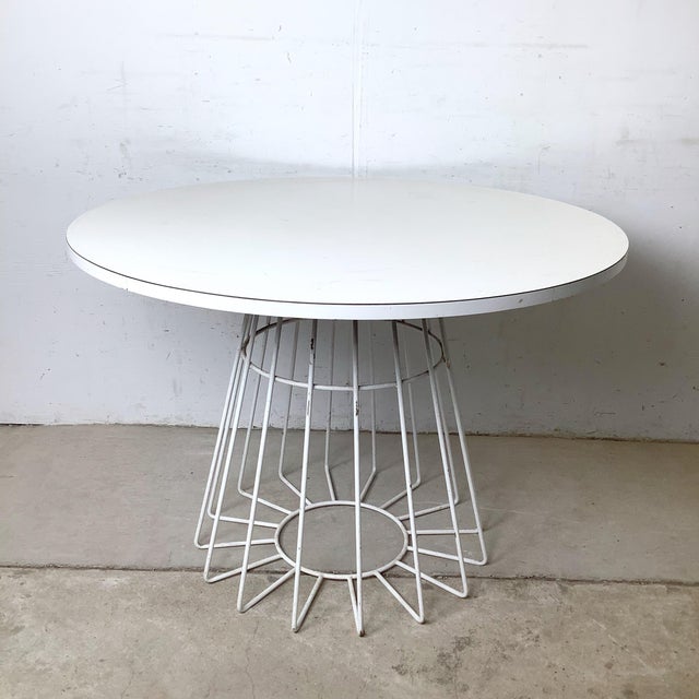 A round white laminate dining table with a Knoll-style wire base — steel rods radiating outward and downward from a...