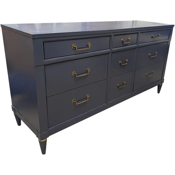 A wide dresser in a deep slate-blue finish with a clean, structured silhouette and tapered legs capped in brass. It...