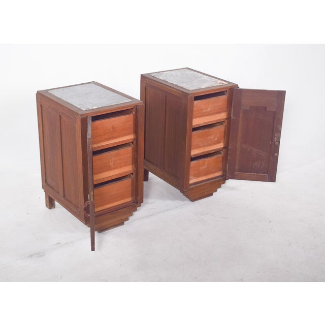 Pair of Deco-style bedside tables. They feature a front door with a simple wooden handle and metal lock, under a carved...