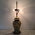 Two-armed table lamp with a base in the shape of a painted vase that features a floral design. The vase is in a vibrant...