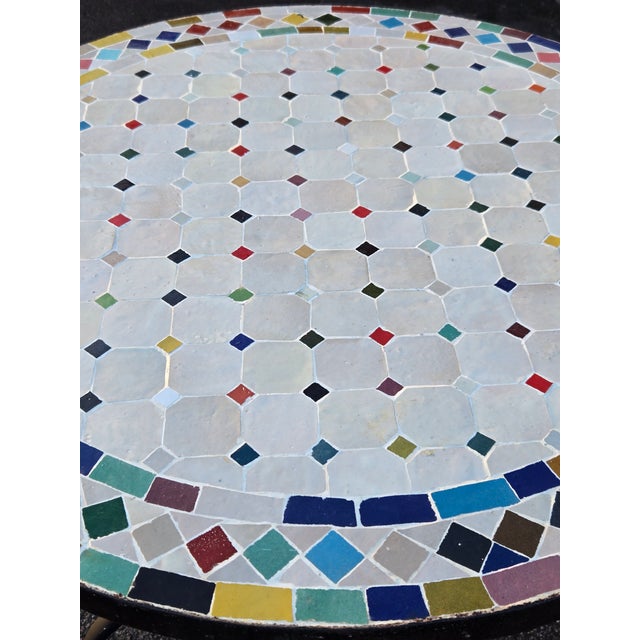 Beautiful round mosaic tile table featuring a vibrant multicolor and off white tiled top and durable black wrought-iron...