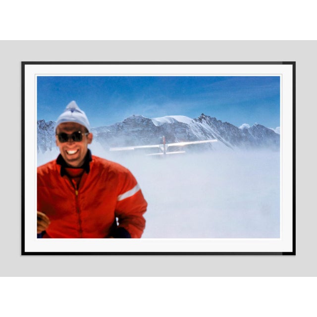 Haute Route Take Off by Toni Frissell A skier smiles from the top of a mountain in the clouds as a plane flies over head,...