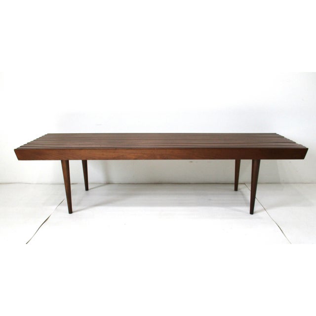A classic Mid Century slat coffee table of bench in the manner of George Nelson in a walnut toned finish and conical legs....