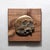 This artwork presents a flat skull rendered in bronze, set against a rich walnut background that enhances its organic...