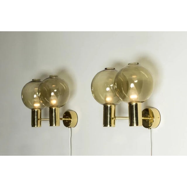 Pair of wall lamps by Hans-Agne Jakobsson, with brass frames and smoke colored glass shades. Sleek brass forms combine...
