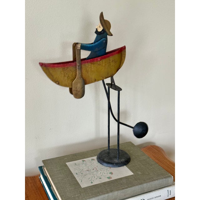A charming vintage folk art sculpture of a man rowing in a rowboat, captured in a kinetic balancing composition. Crafted...
