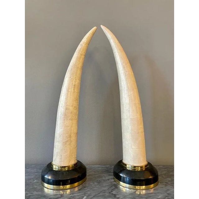A pair of tall faux tusks in tessellated marble mounted on dark marble plinths with brass accents. A tessellation or...
