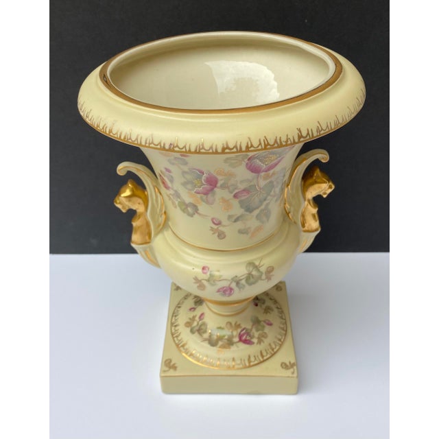 This lovely decorative ceramic urn features embossed accents, hand-painted delicate flowers, and gold griffin handles. The...