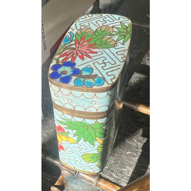 A jewel-like cloisonné box featuring brilliant cobalt blue, sunny yellow, and coral pink flowers against a soft turquoise...