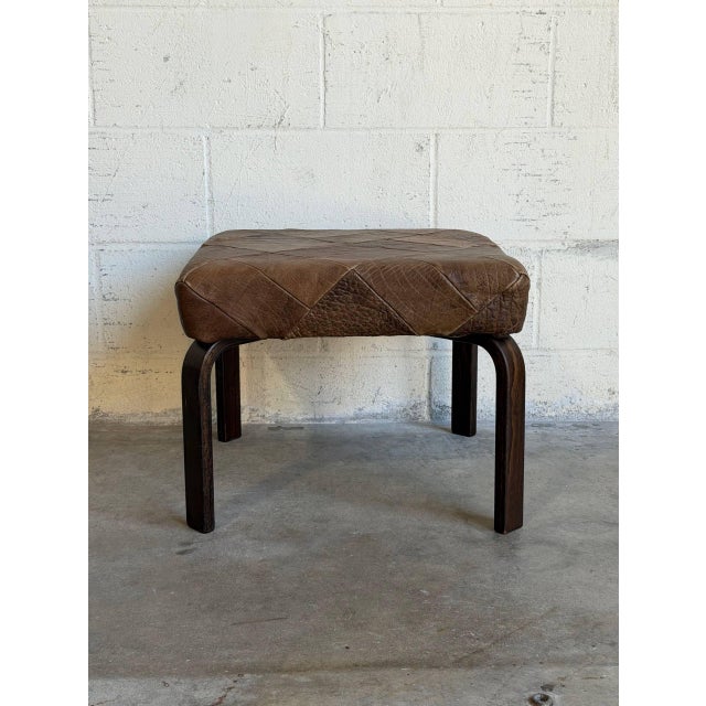 A square patchwork ottoman featuring a cushioned top set over curved bentwood legs. Its minimalist form delivers organic...