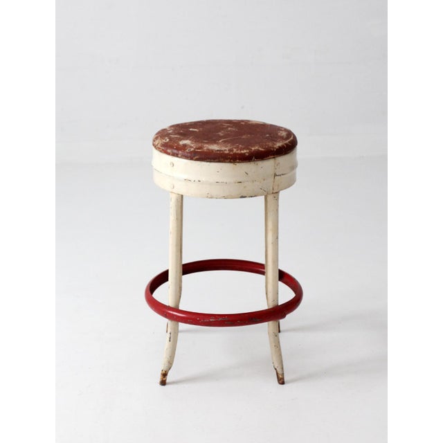 Vintage mid-century industrial stool featuring a cream-painted metal frame, red circular footrest, and upholstered seat....