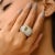 A glamorous Art Deco–inspired statement ring showcasing a radiant emerald-cut green cubic zirconia paired with a brilliant...