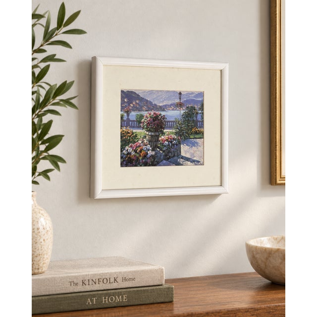 A charming framed wall artwork featuring a serene lakeside garden scene with blooming florals, soft mountain views, and a...