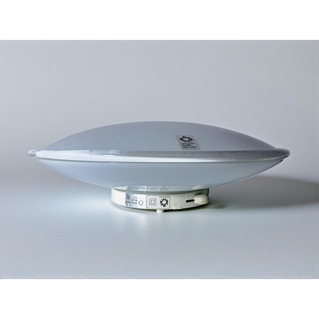 The lamp features a round, flat shape, with a white glass body that ensures soft, even light diffusion, creating a...