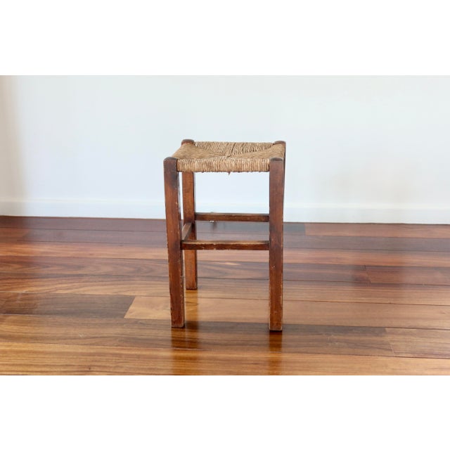 Rustic antique stool with a wooden frame and woven straw seat, typical of utilitarian country furniture. The simple lines...