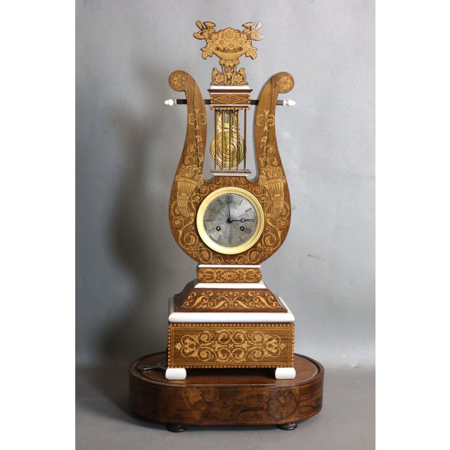 Large lyre-shaped clock in wood veneer and marquetry of light wood, decorated with scrolls, volutes, and cornucopias, the...