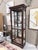 A finely crafted cherry curio cabinet by Howard Miller, presenting a graceful silhouette with classic architectural...