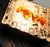 A distinctive wall lamp made of grey wrought iron with a textured finish and delicate filigree glass featuring amber-...
