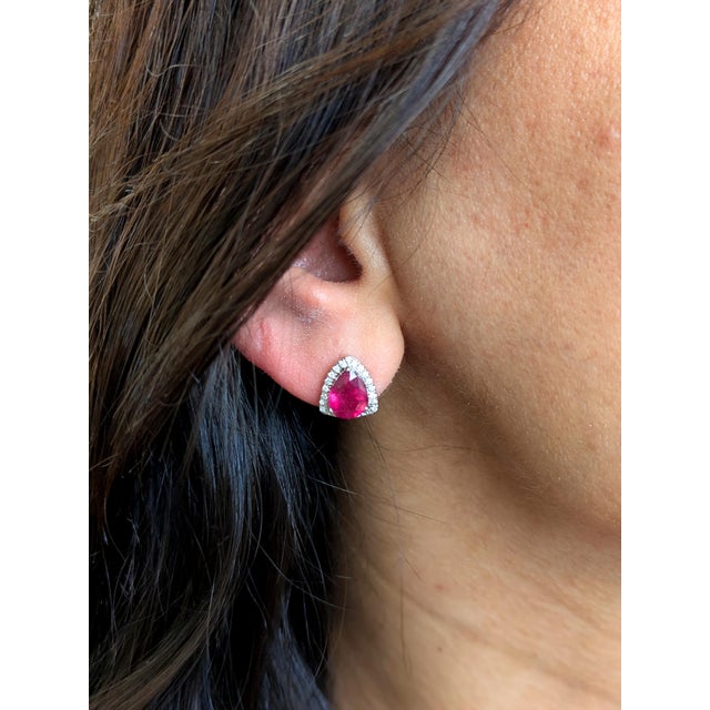These beautiful stud earrings feature a pair of 4.7 carat weight trillion cut ruby gemstones with a halo of natural earth...