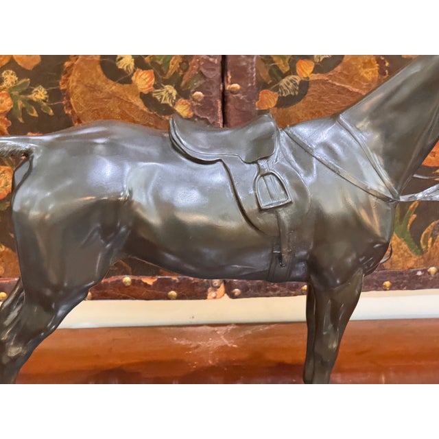 Standing saddled horse in dark patinated cast metal, modeled with bridle, saddle, and stirrups, on a textured rectangular...