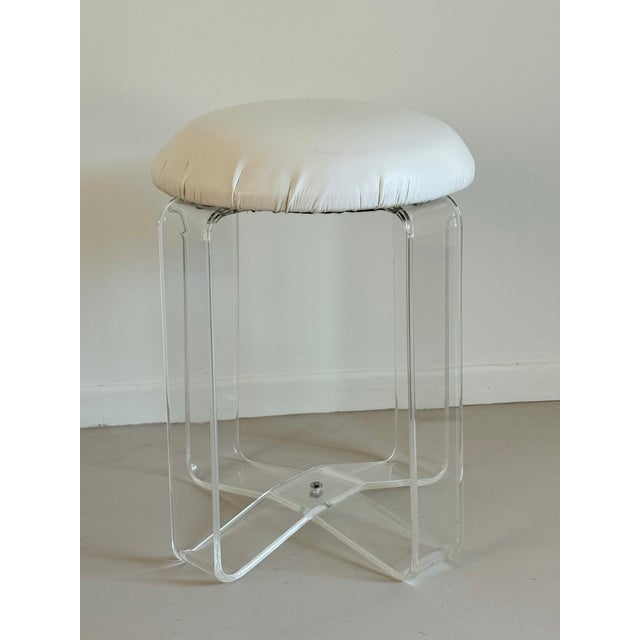 A lucite low stool with vinyl upholstered seat. The ribbon-like clear acrylic legs cross in at the base. The padded round...