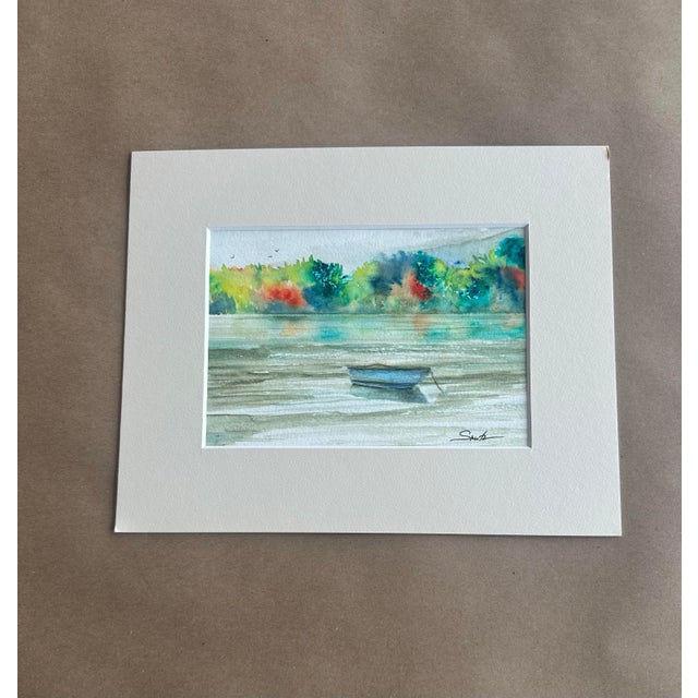 A serene watercolor landscape featuring a solitary boat resting on calm reflective waters. The distant shoreline bursts...