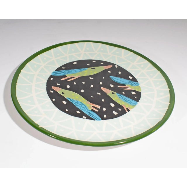 A ceramic bird plate designed by Droll Designs. The center of the plate has four green and blue birds with orange beaks...