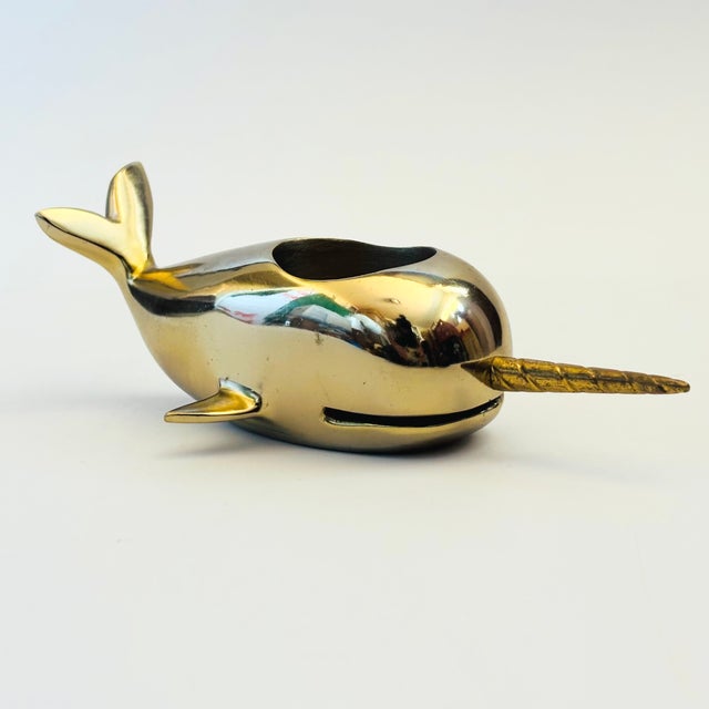 A vintage brass votive candle holder in the shape of a narwhal. The body of the narwhal is a more muted colored gold while...