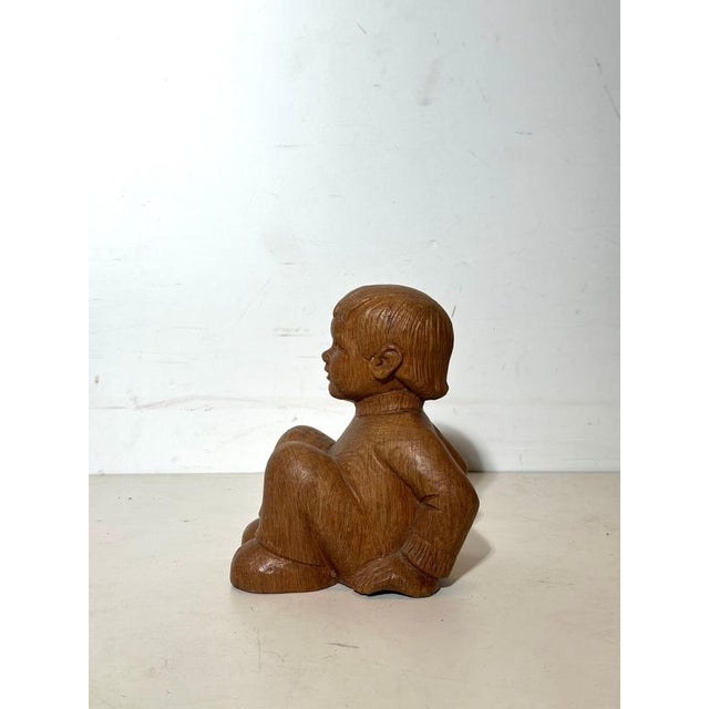 Expressive carved wooden sculpture depicting a seated child, executed with a sober yet deeply emotional presence. The...