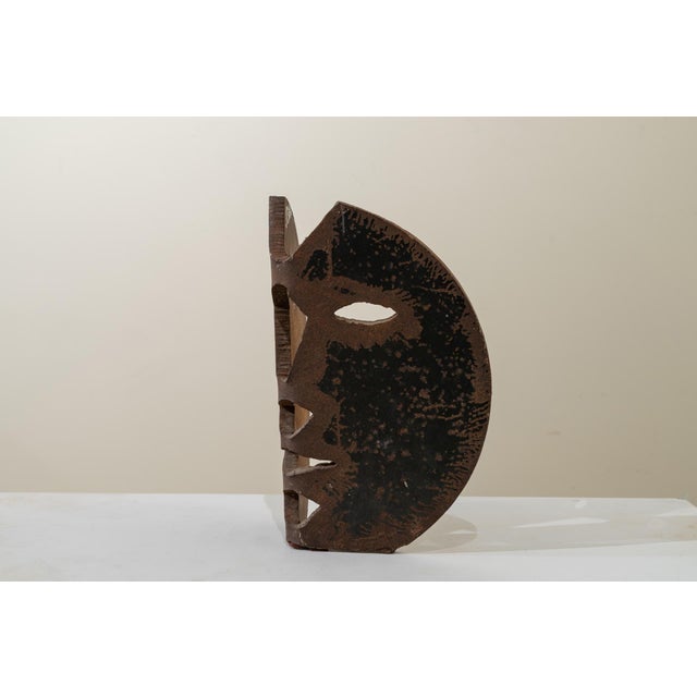 Brutalist mask sculpture made from a thick, concave, oval-shaped iron sheet. The piece features a rough but expressive...