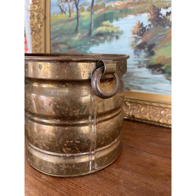 This vintage brass toned bucket features a cylindrical shape with two sturdy side ring handles and horizontal banded...